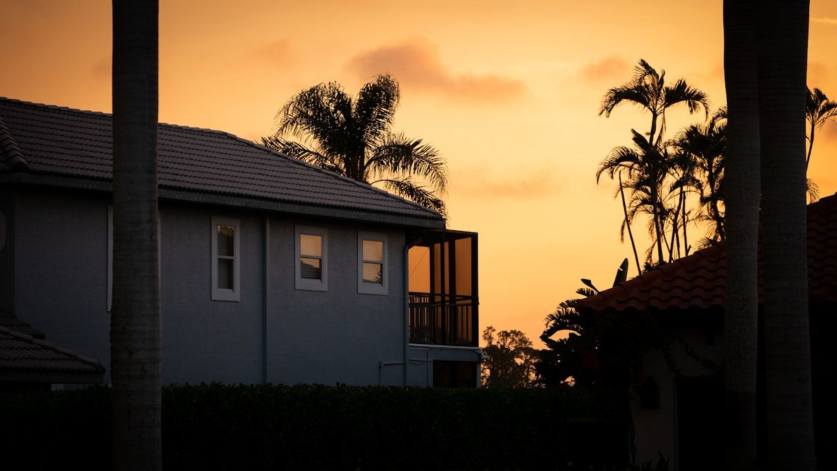 A Florida suburban home viewed from the driveway with an inspector on a ladder examining the roof edge after a recent storm, with scattered palm fronds and damp shingles visible under bright sunlight