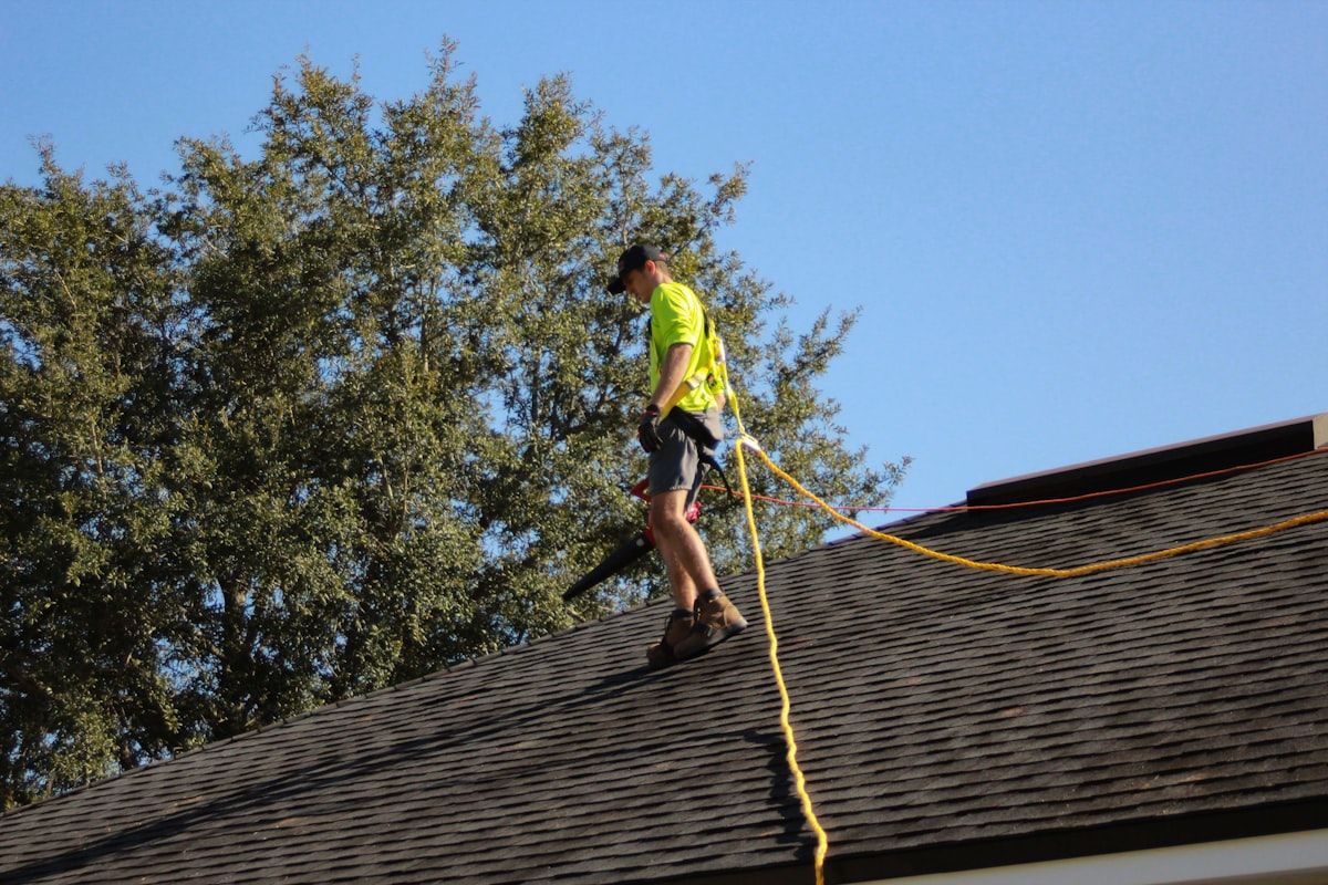 A Southwest Florida home with a roofer on a ladder examining cracked roof tiles and flashing near a vent, while a ceiling stain is visible inside an adjacent room, showing the connection between hidden roof damage and an indoor leak