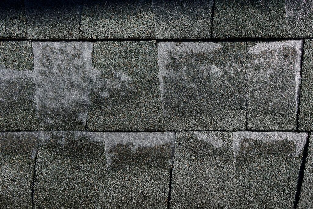 A wide view of an aging shingle roof with curled edges, bare patches of lost granules, a few sagging spots, and multiple areas exposed after a storm, with roofers removing old shingles and exposing sections of the roof deck beneath.