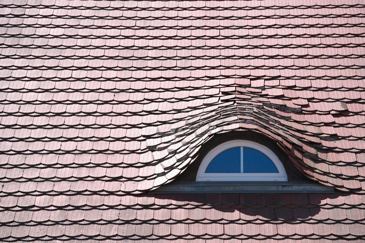 A detailed roof inspection in progress with a technician on the roof photographing cracked tiles and worn flashing, while a second view shows the attic below with exposed rafters, damp insulation, and a flashlight beam highlighting a stain on the underside of the roof decking
